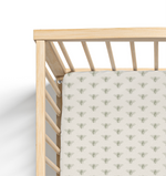 A crib sheet with a sage bee pattern on a light beige background, fitted on a crib with wooden sides.