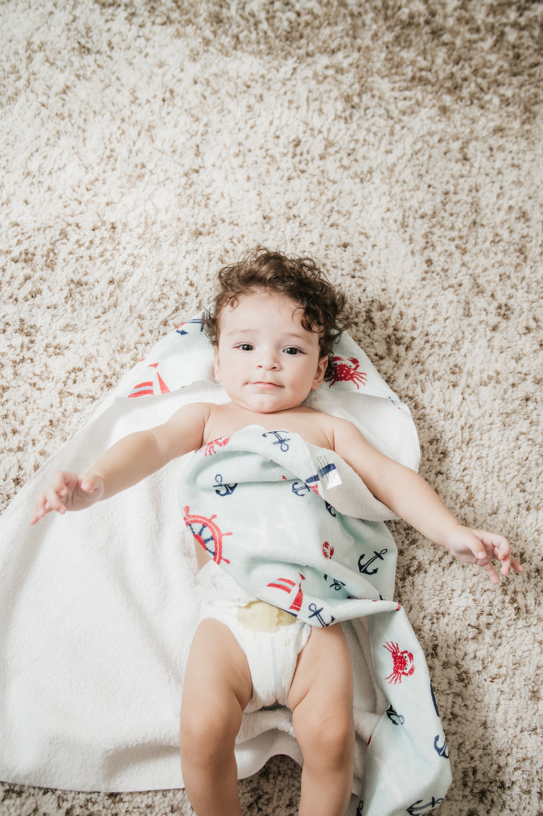 Baby lying on a hooded baby towel with a pattern of anchors and crabs on a beige carpet.