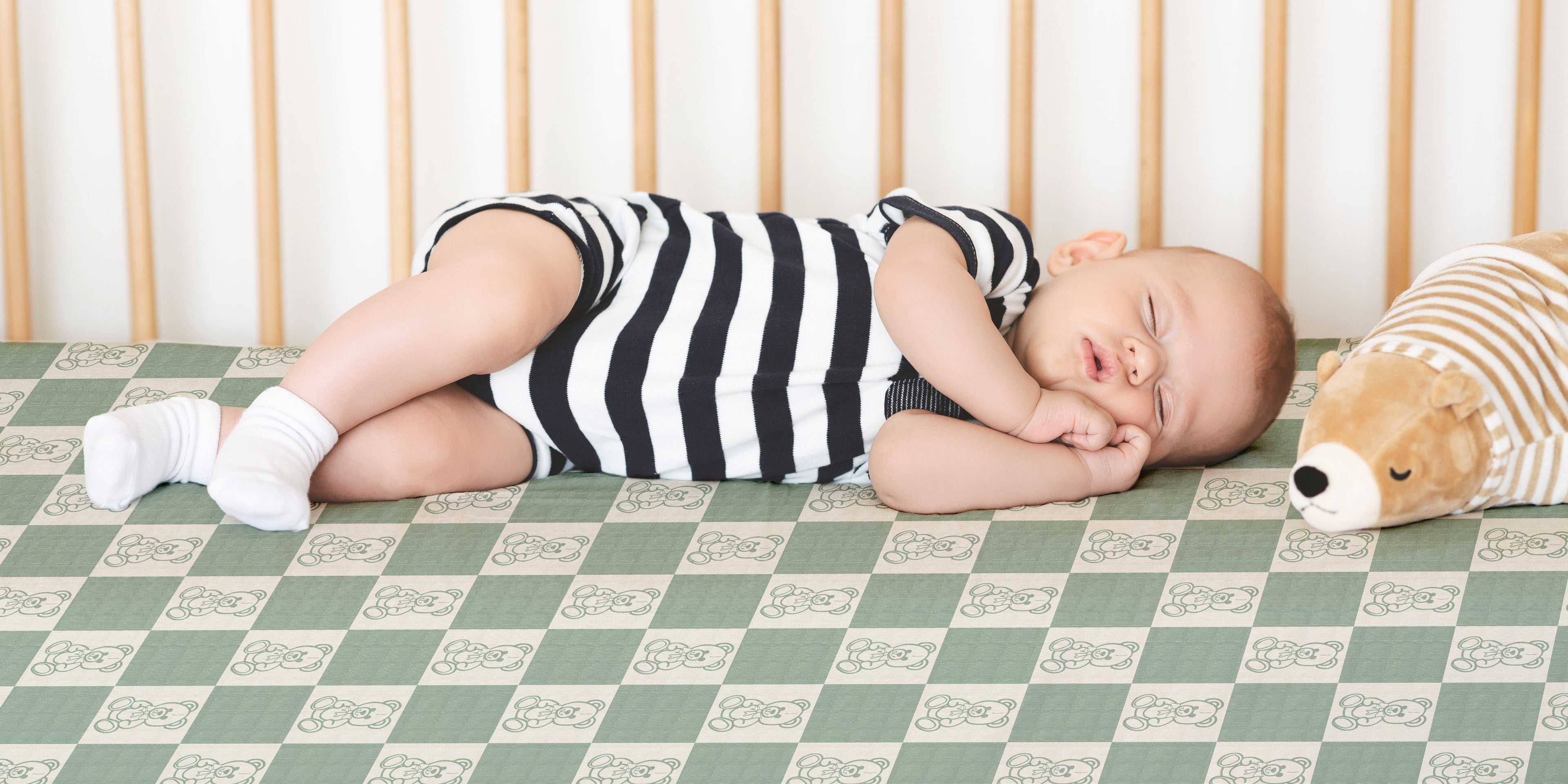 Baby sleeping in a crib with a checkered bears crib sheet in a striped outfit and teddy bear.